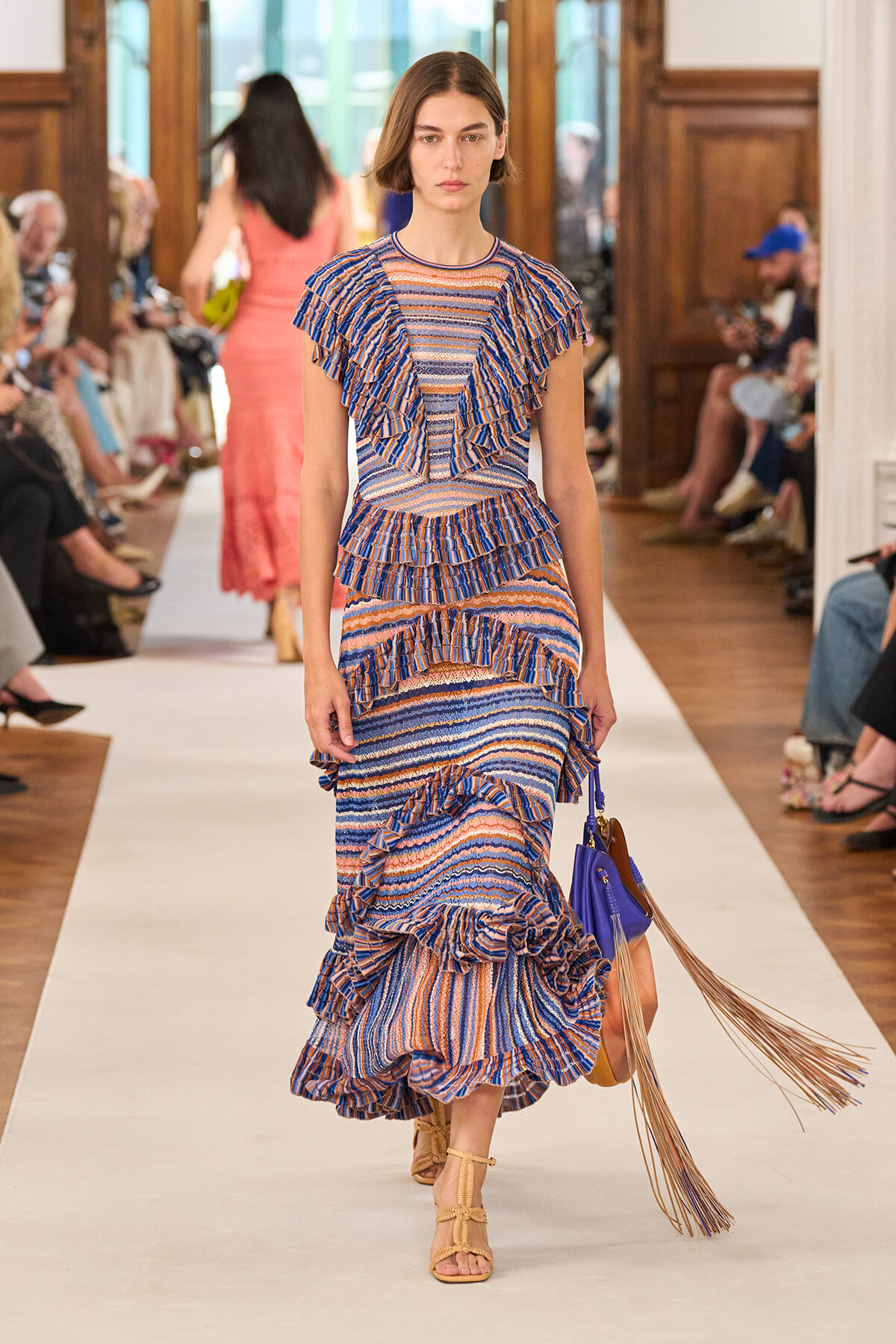 Model walking the runway in a blue-and-orange striped, layered ruffle dress with a purple handbag and tan sandals; audience seated along the catwalk in a fashion show venue.