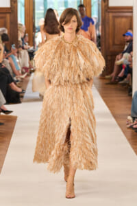 Model walking the runway in a beige feathered evening gown with a short cape, gold necklace, and strappy sandals.