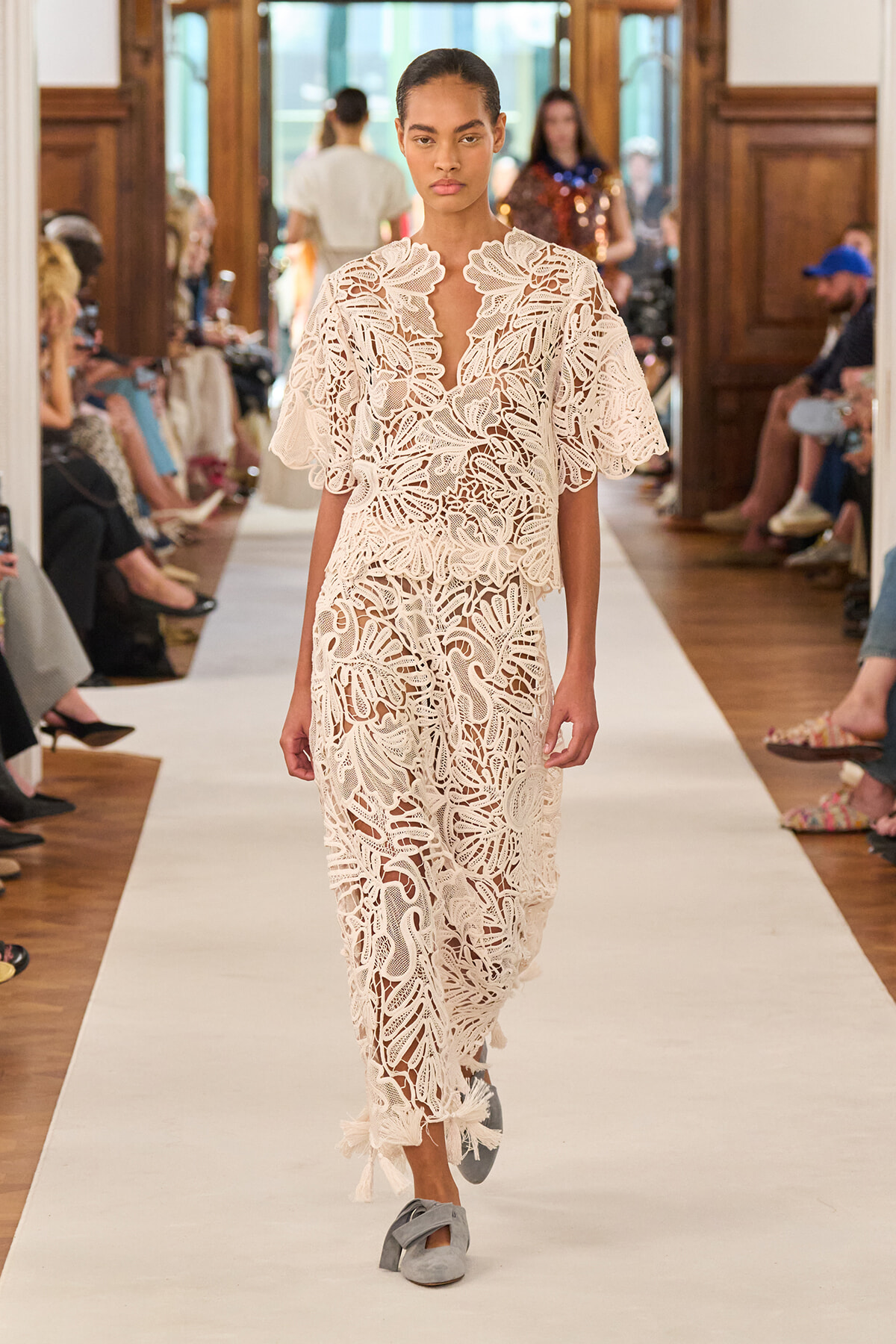 Model walks a runway in an ivory lace dress with a bold floral pattern and a cape-like overlay, audience seated on either side.