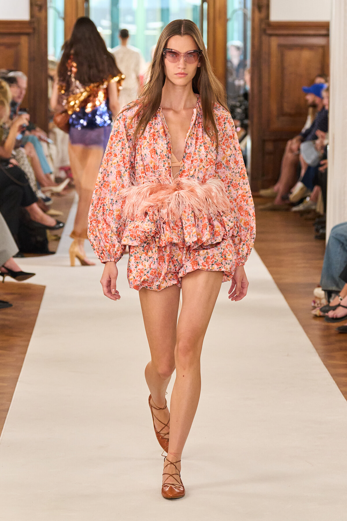 Model walking a fashion runway in a pink floral romper with a feathered waist belt and pink-tinted sunglasses.