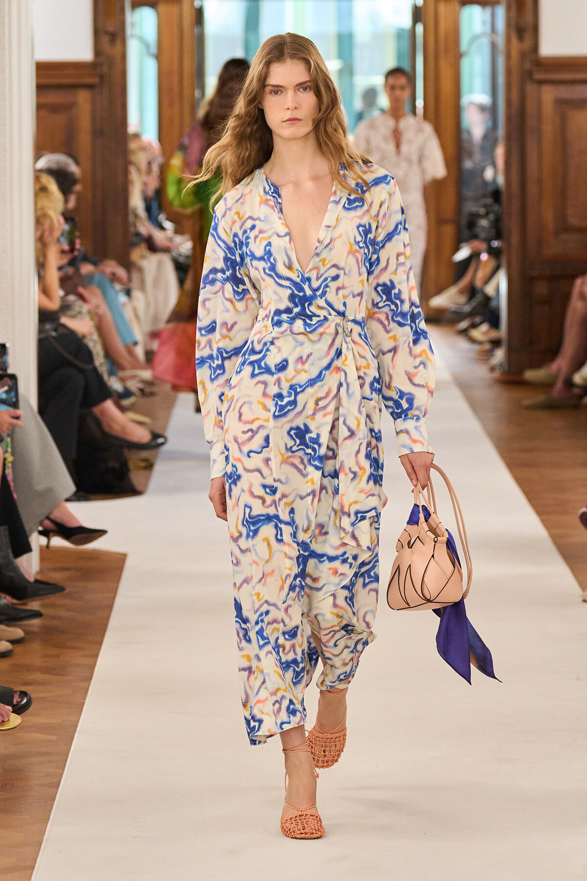 Model walks a fashion runway in a blue, white, and yellow abstract-wrap dress, carrying a pink handbag with a purple scarf.