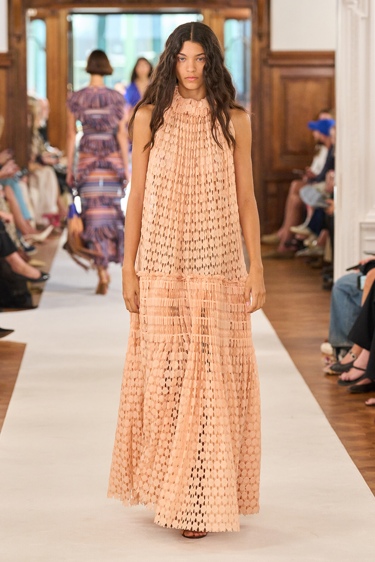 Model walks the runway in a peach sleeveless maxi dress with circular cutouts and a ruffled lower tier.