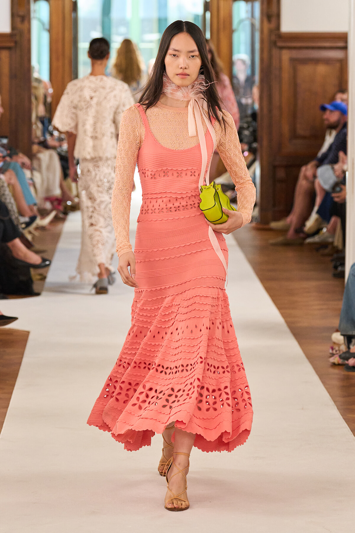 Model walking a runway in a coral pink lace dress with long sleeves, a scarf around the neck, and a neon yellow-green small bag.