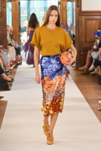 Model walking a fashion show wearing a mustard crop top and a gradient sequin skirt, clutch in hand.