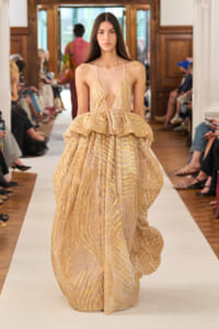 Model walks the runway in a champagne-gold gown with metallic stripes and a dramatic, voluminous ruffled skirt.