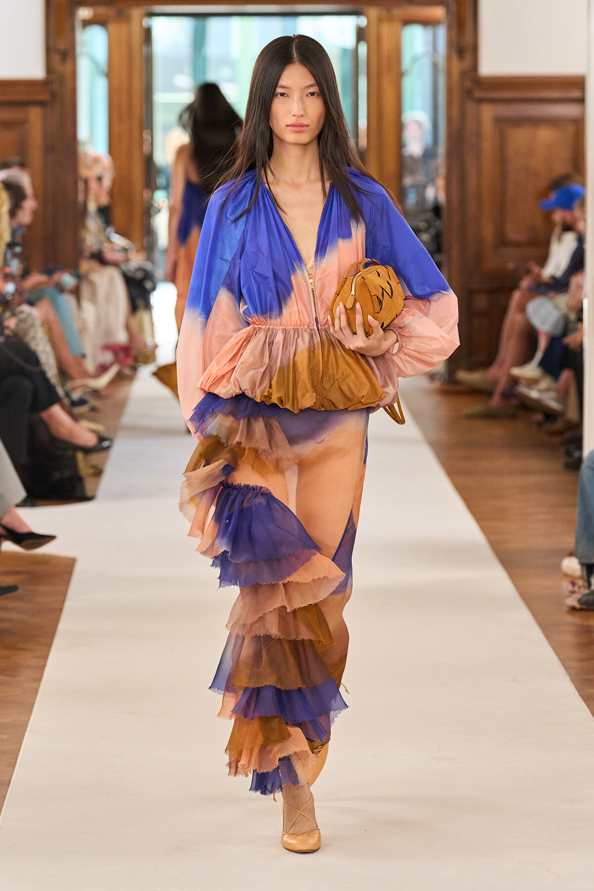 Model walking the runway in a blue, peach, and brown tiered tulle dress, holding a tan clutch.