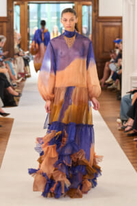 Model walks a fashion runway in a sheer, color-block gown with a ruffled, layered train in peach, purple, and blue tones, accessorized with a beaded necklace.