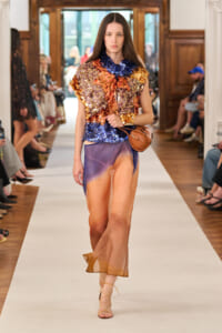 Model walks runway in a multicolored sequined top paired with a sheer gradient orange-to-purple skirt, tan sandals, and a brown crossbody bag.