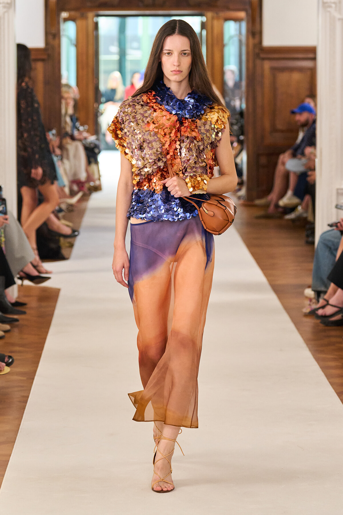 Model walks runway in a multicolored sequined top paired with a sheer gradient orange-to-purple skirt, tan sandals, and a brown crossbody bag.