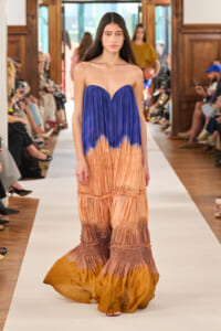 Model walking a runway in a strapless ombre maxi dress fading from cobalt blue to peach-brown with ruched tiers, audience along the sides.