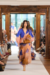Model walks down a fashion runway in a blue-pink-tan layered dress, holding a brown clutch as audience watches from sides in a grand wooden setting.