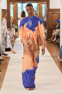 Model walks the runway in a peach-and-blue tie-dye maxi outfit with draped ruffles at a fashion show.
