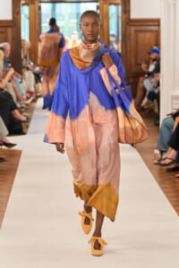 Model walks a runway in a color-blocked blue-to-peach oversized robe with scarf and matching tote; audience sits along the catwalk.