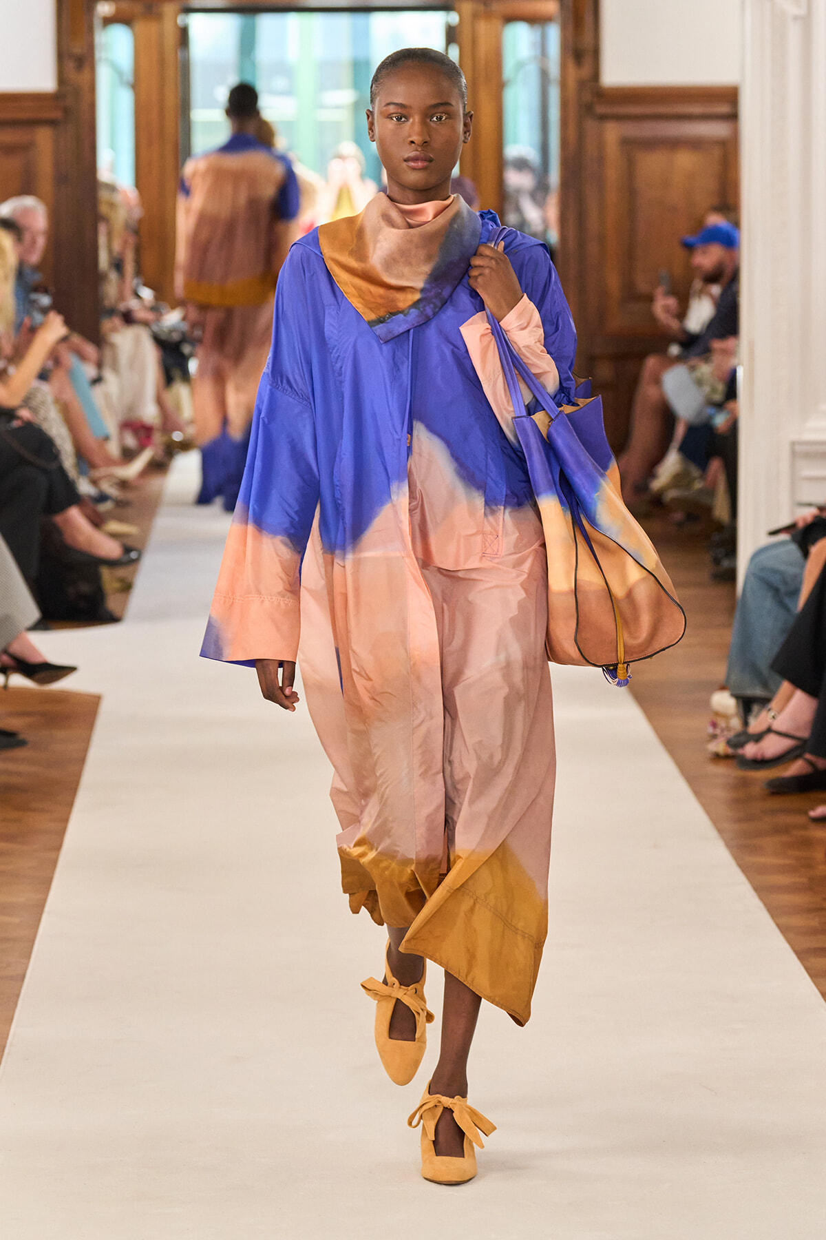 Model walks a runway in a color-blocked blue-to-peach oversized robe with scarf and matching tote; audience sits along the catwalk.
