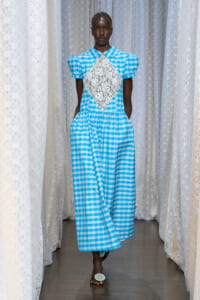 Model wearing a blue gingham dress with a white lace diamond panel at the chest, standing between lace-curtained walls.
