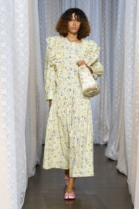 Model wearing a pale yellow, long-sleeve dress with ruffles and a cat-print pattern, carrying a matching shoulder bag, standing among lace curtains.
