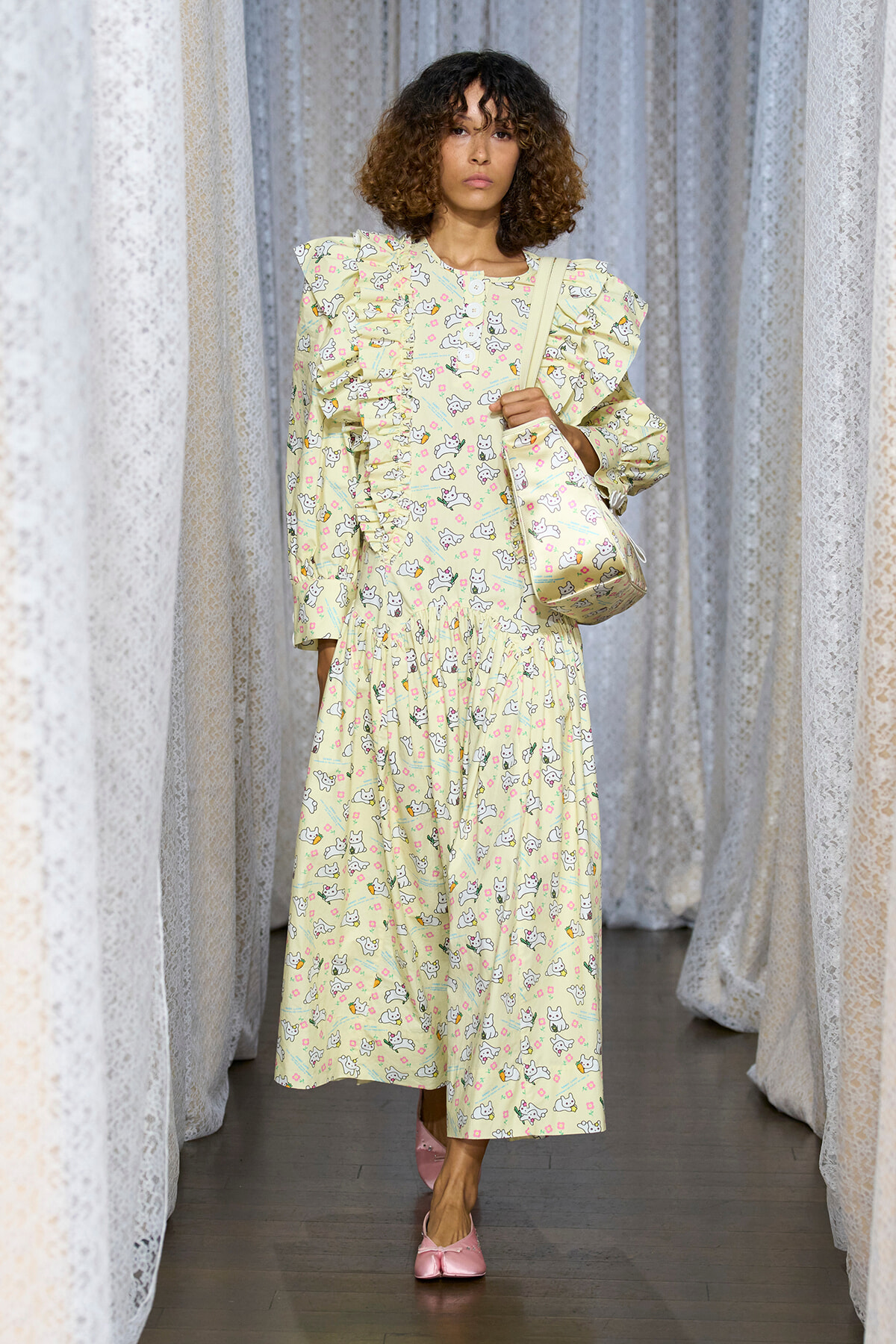 Model wearing a pale yellow, long-sleeve dress with ruffles and a cat-print pattern, carrying a matching shoulder bag, standing among lace curtains.