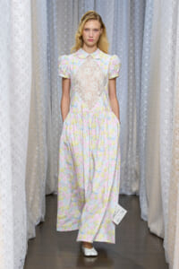 Female model on a runway wearing a pastel floral dress with lace bodice and short sleeves.