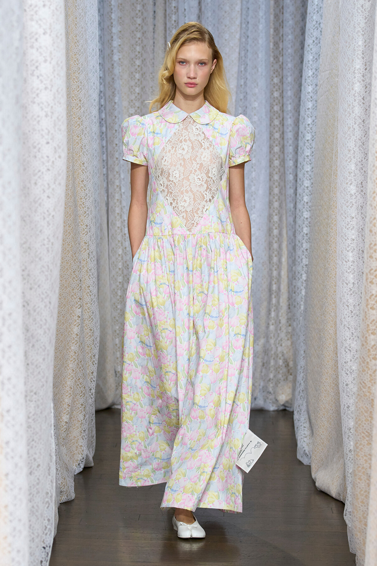 Female model on a runway wearing a pastel floral dress with lace bodice and short sleeves.