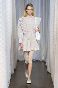 Model walking a runway in a pastel floral dress with ruffles, lace leggings, white flats, and a white shoulder bag.