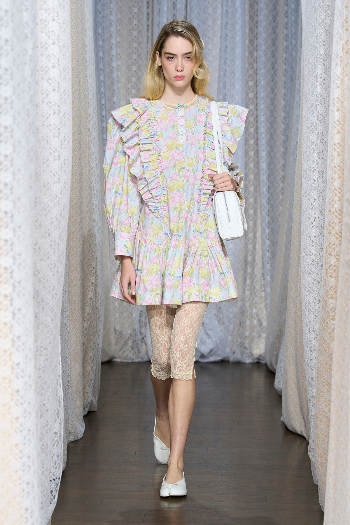 Model walking a runway in a pastel floral dress with ruffles, lace leggings, white flats, and a white shoulder bag.