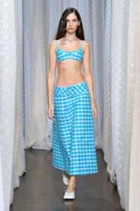 Model walking a fashion show wearing a blue plaid bikini top and matching long skirt, white flat shoes, against a backdrop of pale curtains.