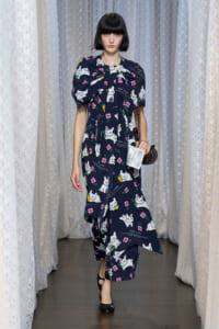 Model walks a runway wearing a navy dress covered in white cat prints and pink flowers, carrying a small brown handbag.