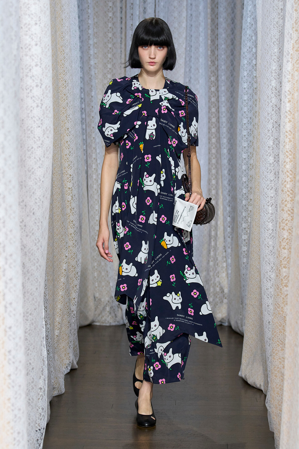 Model walks a runway wearing a navy dress covered in white cat prints and pink flowers, carrying a small brown handbag.