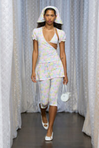 Model walking a runway in a pastel floral wrap-top and capri pants set with matching hat, carrying a small white circular handbag.