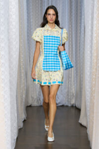 Model walking a runway in a pale yellow printed dress with puff sleeves, a bright blue gingham bib, and a matching blue tote bag.