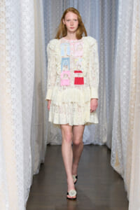 Model walking the runway in a cream lace dress with ruffled details and pastel patchwork panels, set against lace curtains backdrop.
