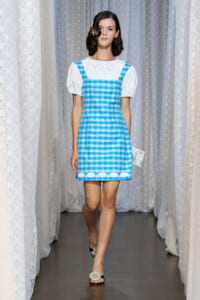 Model walking a runway in a blue-and-white gingham pinafore over a white puff-sleeve blouse.