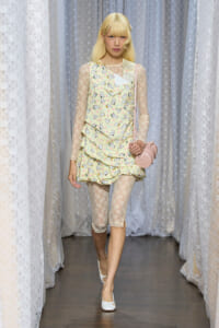 Model walking a runway in a pale green dress with a small animal print and lace sleeves, carrying a pink round bag