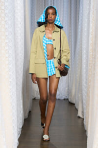 Model walking a fashion runway in a blue plaid bikini top and shorts under a tan oversized jacket, with a blue plaid headscarf and a brown crossbody bag.