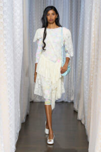 Model walking the runway in a pastel floral lace dress with a tiered, cream skirt and a light blue crossbody bag nearby shoulder height.