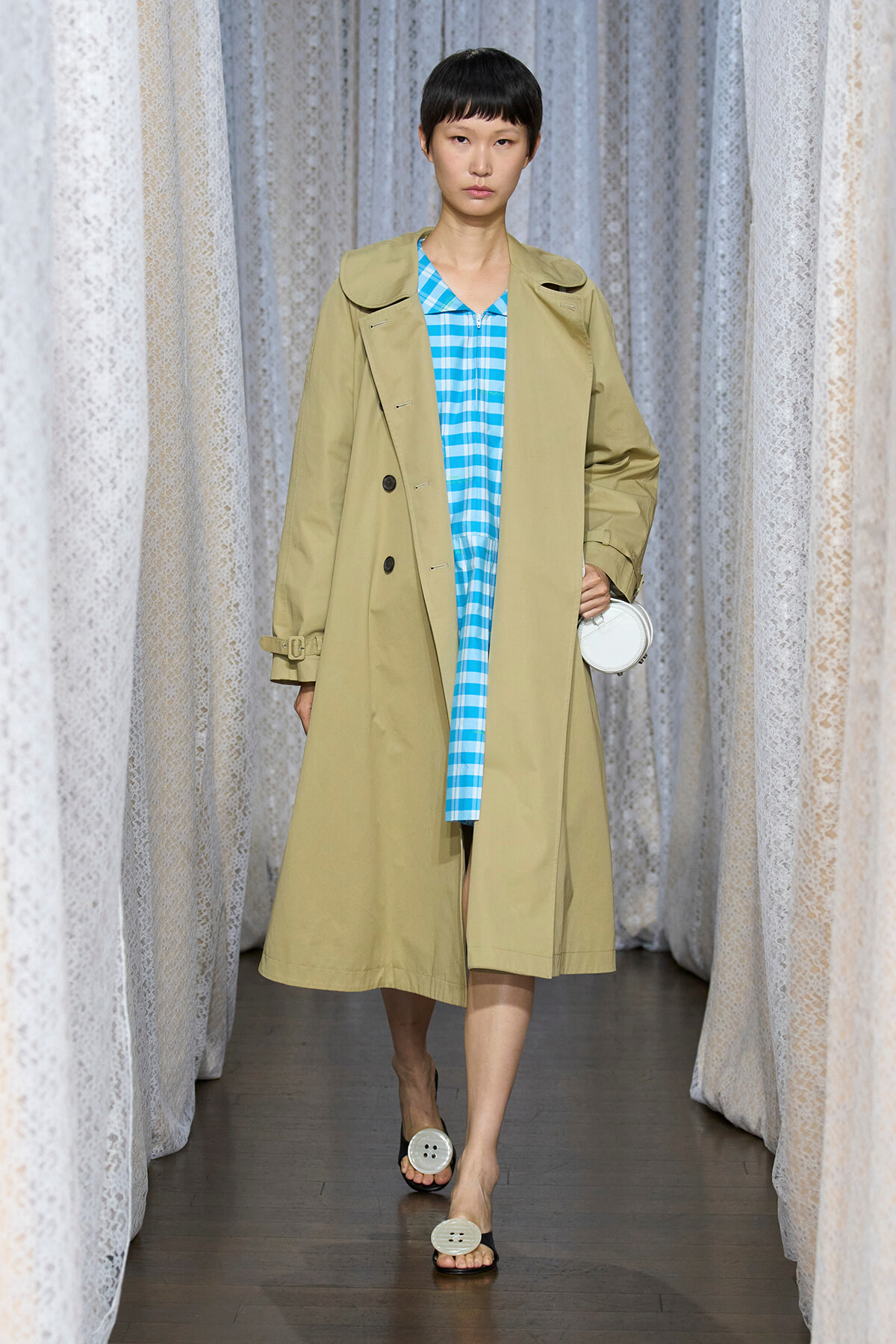 Runway model wearing a beige trench coat over a blue gingham dress, carrying a small white round bag.