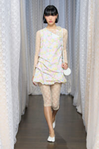 Model walking the runway in a pastel floral layered dress with sheer lace sleeves, beige lace leggings, white flats, and a small white purse.