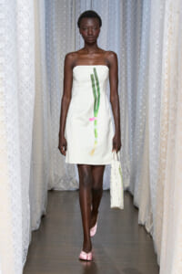 Model walks a runway wearing a white strap dress with a green vertical design, carrying a white speckled tote and wearing pink flats.