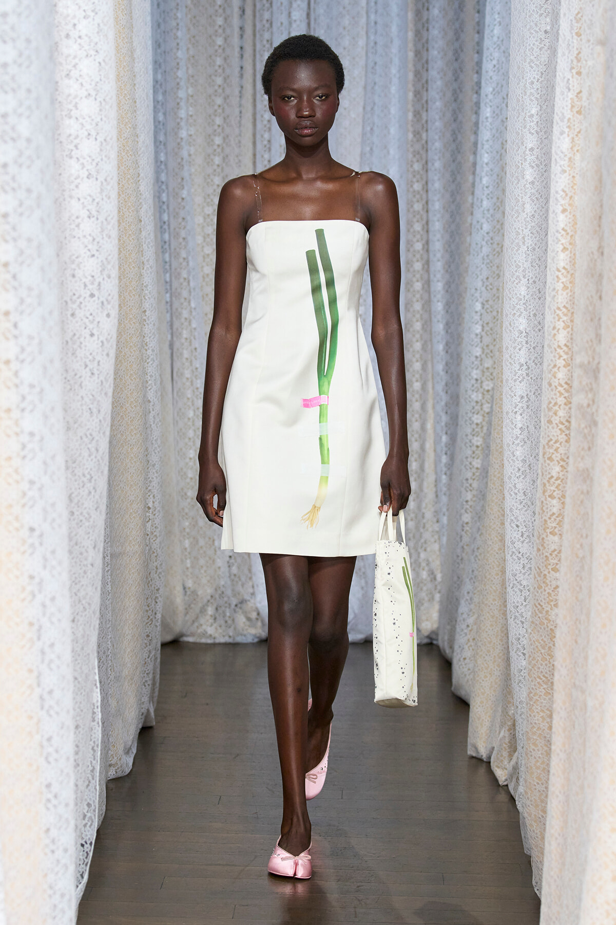 Model walks a runway wearing a white strap dress with a green vertical design, carrying a white speckled tote and wearing pink flats.