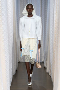 Model walking a runway in a white Victorian-inspired outfit: lace veil, puffed-sleeve blouse, lace skirt with blue ribbons, white tights, white heels, and a pink handbag