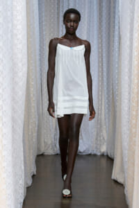 Model walking a runway in a white slip dress with a small blue bow, lace backdrop.