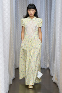 Model walking the runway in a pale yellow maxi dress with a lace front panel and a playful cat print, hands in pockets, lace-curtained backdrop.
