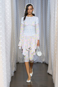 Model on a fashion runway wearing a pastel floral layered dress with long sleeves, headscarf, white heels, and a small white handbag.