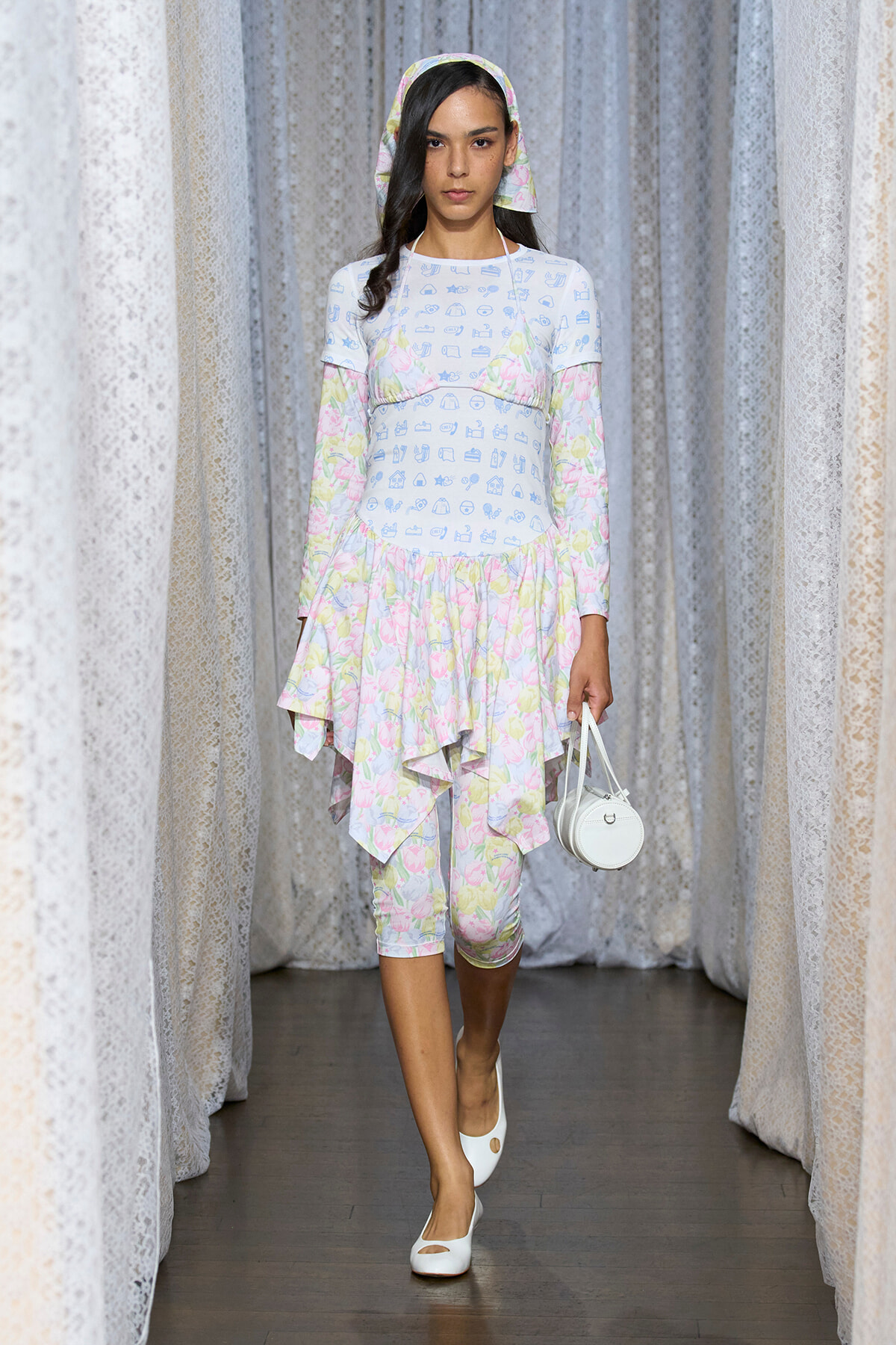 Model on a fashion runway wearing a pastel floral layered dress with long sleeves, headscarf, white heels, and a small white handbag.