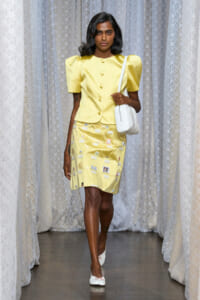 Model walks a runway in a pale yellow dress with puff sleeves, large buttons, and a sticker-covered skirt, carrying a white handbag and wearing white flats.