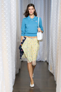 Model walking a runway in a light blue cropped polo sweater, a pastel cat‑print wrap skirt, white handbag and white flats.