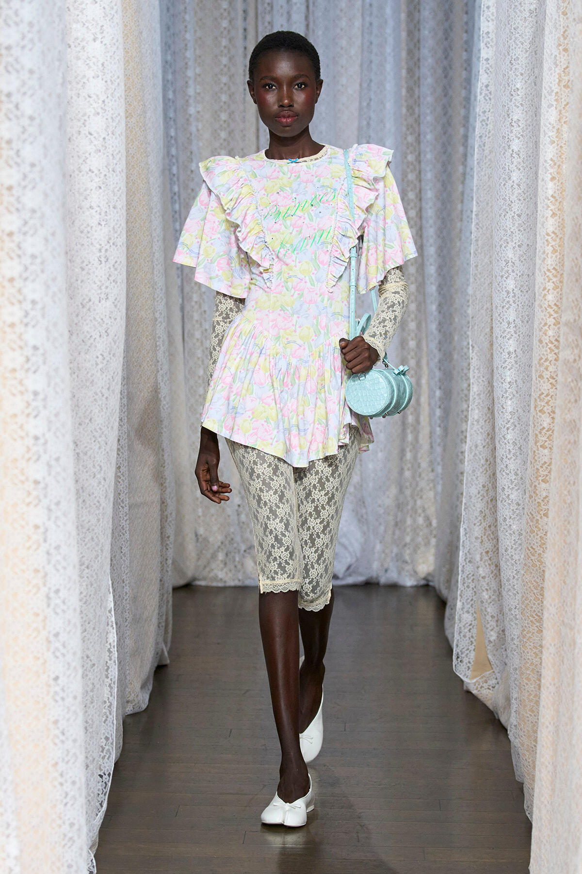 Model walking the runway in a pastel pink, blue, and yellow floral tunic with ruffled sleeves paired with cream lace capris and white flats, carrying a light blue round handbag.