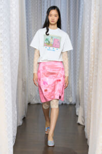 Model walks a runway wearing a white graphic T-shirt, pink satin skirt, and lace sleeves with matching lace leggings and pastel flats.