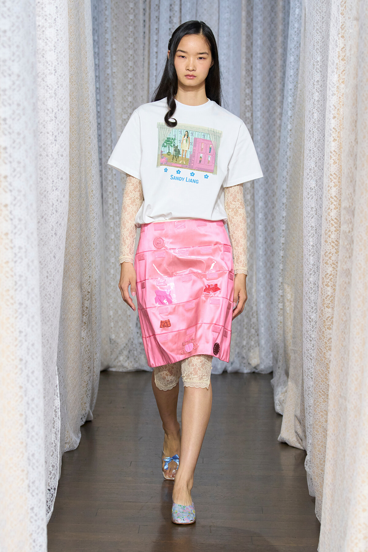 Model walks a runway wearing a white graphic T-shirt, pink satin skirt, and lace sleeves with matching lace leggings and pastel flats.