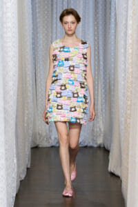 Model walking a fashion runway in a pastel multicolor patchwork ruffle dress with pink flats against lace curtains backdrop.